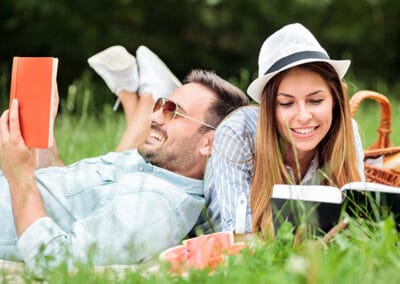 A smiling couple relaxes on a picnic blanket in the spring sunshine, reading and laughing together. A wicker picnic basket and soft grass complete the tranquil outdoor setting.