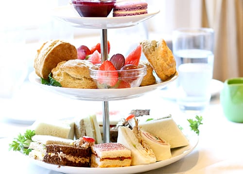 A traditional afternoon tea set in a garden with fine china teacups, finger sandwiches, scones with jam and cream, and dainty cakes arranged on a three-tiered stand.