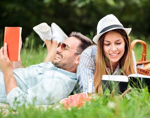 Happy young couple enjoying a good read during picnic in a park A smiling couple relaxes on a picnic blanket in the spring sunshine, reading and laughing together. A wicker picnic basket and soft grass complete the tranquil outdoor setting.